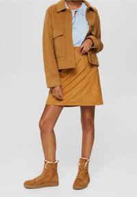 Femme portant une veste couleur camel, une jupe assortie, un chemisier bleu clair et des bottines à lacets beige avec doublure en fourrure, debout devant un fond gris.