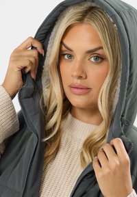 Gilet rembourré gris avec une capuche doublée, pull en tricot beige texturé en dessous, et fermeture zippée. Le modèle tient la capuche du gilet.