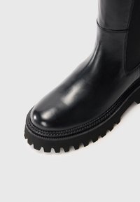 Botte en cuir noir avec semelle à crampons épaisse et bout arrondi, dotée d'un panneau latéral en élasthanne et de coutures détaillées sur fond blanc.