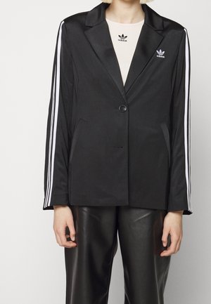 Osoba nosi czarną marynarkę Adidas z białymi paskami na rękawach, czarne skórzane spodnie oraz białą koszulkę Adidas widoczną przy kołnierzu.
