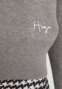 Szary sweter dzianinowy z ribbrowym wykończeniem, z różowym haftowanym logo "Hugo". W zestawie z spódnicą w czarno-białą kratę w stylu houndstooth.