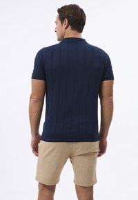 Marineblaues Poloshirt mit kurzen Ärmeln, strukturierten vertikalen Mustern, geripptem Kragen und Saum. Kombiniert mit beigen Leinen-Shorts.