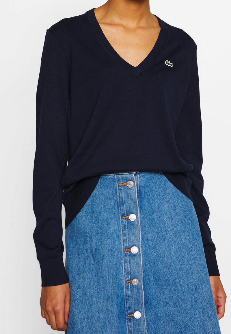 Femme portant un pull bleu marine à col en V et manches longues avec un petit logo brodé, et une jupe en jean bleu taille haute avec boutons sur le devant.