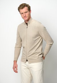 Suéter beige con cremallera, hecho de una tela suave, con cuello alto y mangas largas, combinado con pantalones de color claro.