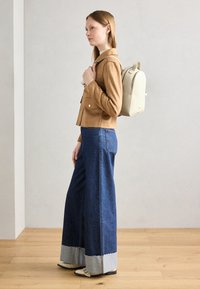 Sveten jakke i lys semsket skinn med to frontlommer, kombinert med vide mørke denimjeans med lys blå oppbrett. Off-white ryggsekk og beige støvler.