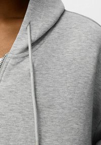 Sudadera con capucha gris con cremallera, confeccionada con una tela suave y texturizada. Cuenta con una capucha grande, cremallera frontal y cordones en un tono más claro.