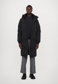 Abrigo acolchado negro con un dobladillo largo, capucha y cierre delantero con cremallera. Cuenta con puños elásticos y dos bolsillos frontales. Llevado sobre pantalones grises y zapatos negros.