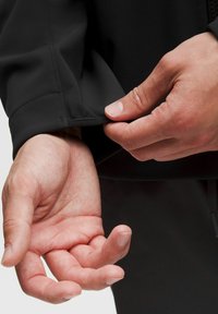 Juodas švarko audinys su lygiu paviršiumi, rodomas artimas vaizdas, kaip ranka reguliuoja rankogalių. Aiškiai matomi siuvimo detalės.