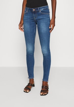 Kobieta w niebieskich obcisłych dżinsach i czarnych sandałach z odkrytymi palcami na słupku, stojąca na jasnym, jednolitym tle.