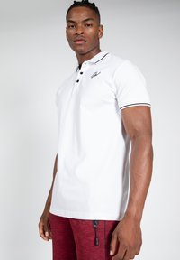 Witte polo met een zwart logo, drie-knopen placket, zwarte trim op de mouwen, gedragen met rode textuur atletische broek met ritszakken.