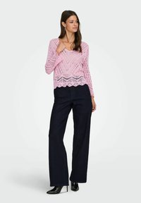 Femme portant un haut à manches longues en crochet rose, un jean large foncé et des bottes à talons hauts noires pointues, debout avec une main posée sur la poitrine.