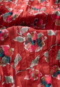 Tessuto floreale in rosso corallo con grandi fiori rosa pallido e blu, accompagnati da foglie verdi. La texture sgualcita aggiunge profondità al design.