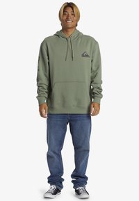 Felpa verde in cotone con tasca frontale, coulisse regolabile e logo. Abbinata a jeans blu e sneakers in bianco e nero.