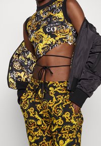 Svart bomberjacka, croptop med guld- och svart mönster, och matchande byxor med intrikata gyllene spiraler på svart bakgrund.