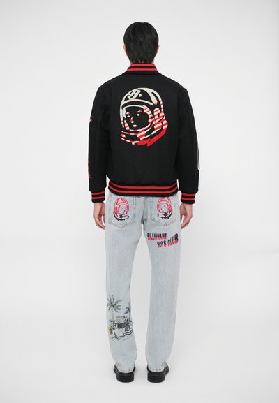 A jaqueta bomber preta apresenta um gráfico de astronauta bordado em destaque nas costas; jeans claros com designs e texto coloridos.