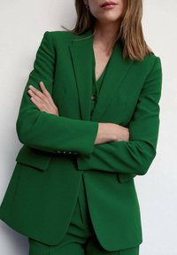 Femme debout, les bras croisés, portant un blazer vert cintré et un pantalon assorti, sur un fond clair uni.