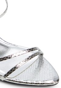 Hopeanvärinen metallinen nilkkaketjullinen kenkä, jossa on terävä kärki, kuviollinen pinta ja ristikkäisnauhamalli. Sileä, kiiltävä pinta.
