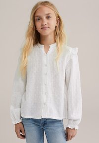 Junge Frau mit langen blonden Haaren, die eine strukturierte weiße Bluse mit Knöpfen und Rüschen an den Ärmeln sowie hellblauen Jeans trägt, vor einem neutralen Hintergrund.