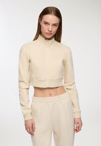 Felpa beige corta con zip e colletto alto, abbinata a pantaloni elasticizzati con vita alta. Tessuto morbido.
