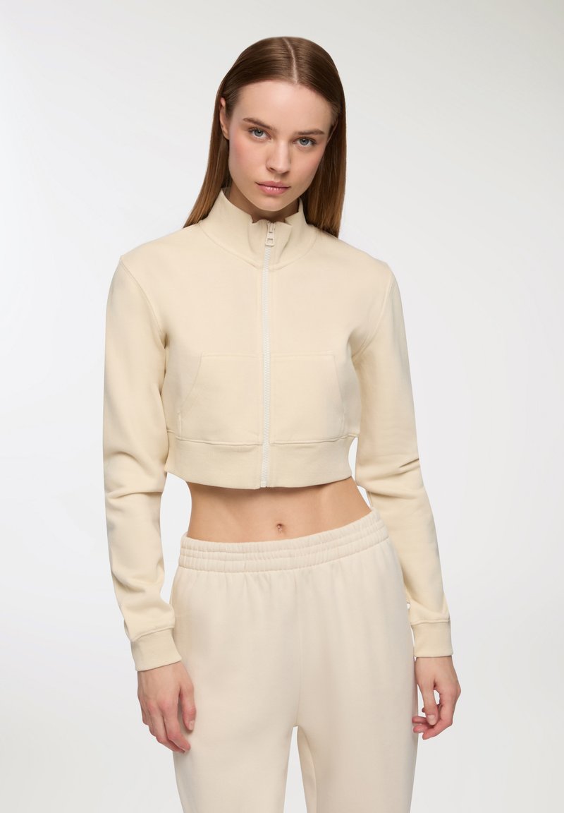 Felpa beige corta con zip e colletto alto, abbinata a pantaloni elasticizzati con vita alta. Tessuto morbido.