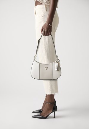 Personne portant un pantalon crème et des talons hauts noirs, tenant un sac à main de créateur blanc avec une bandoulière noire et un logo en métal.