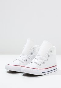 Białe trampki typu high-top wykonane z płótna, z sznurowaniami, srebrnymi oczkami, gumowymi podeszwami oraz czerwonymi i niebieskimi paskami wzdłuż spodu.