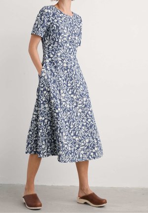 Femme portant une robe midi à manches courtes bleue et blanche à motifs floraux avec des poches et des sabots en bois marron, debout contre un mur clair uni.