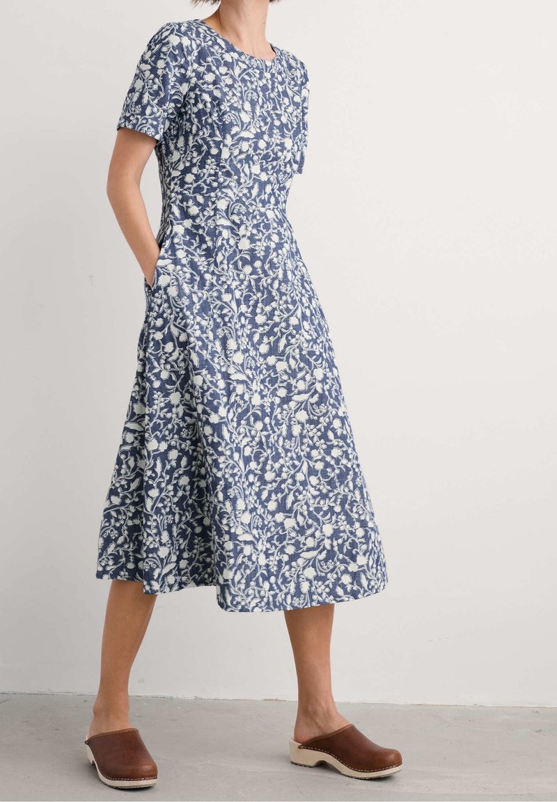 Femme portant une robe midi à manches courtes bleue et blanche à motifs floraux avec des poches et des sabots en bois marron, debout contre un mur clair uni.