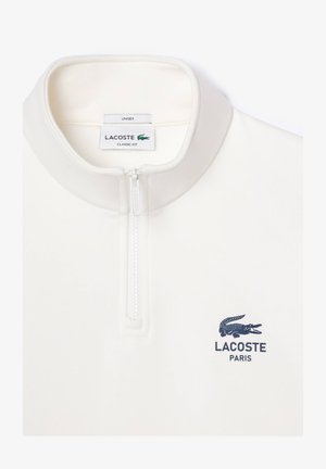 Pullover branco com fecho zip e um corte clássico, apresentando um pequeno logotipo e etiqueta Lacoste em azul marinho. Textura suave e design de gola alta.
