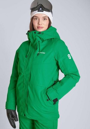 Nainen pukeutuneena vihreään laskettelutakkiin ja -housuihin, valkoinen pipo ja laskettelulasit, sekä mustat hansikkaat, seisomassa vaalean harmaan taustan edessä.