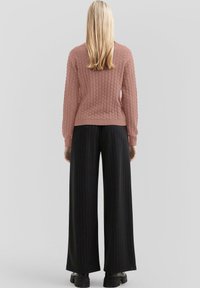 Teksturowany różowy sweter w wzór plastra miodu, zestawiony z czarnymi spodniami w paski o wysokim stanie i szerokich nogawkach. Widoczne czarne buty na platformie.