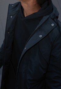 Doudoune marine foncé avec capuche, dotée d'un zip à l'avant et d'une fermeture à pression, fabriquée en tissu lisse et résistant à l'eau.