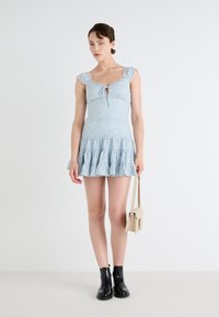 Svijetloplava čipkana haljina s volančićima, raširenim suknjom i otvorom, u kombinaciji s malom bež torbicom i crnim gležnjačama.