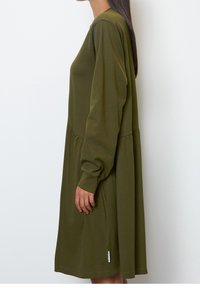 Zielona oliwkowa sukienka z długimi rękawami, dopasowanym gorsetem i rozkloszowanym dołem. Wykonana z miękkiego materiału o delikatnym blasku i z bocznym akcentem w postaci metki.