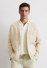 Helles beige Leinenblazer mit einer Brusttasche und zwei Fronttaschen, vorne geknöpft, kombiniert mit einem strukturierten hellen Pullover.