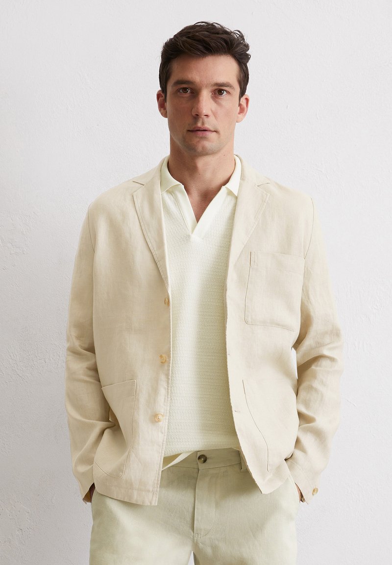 Helles beige Leinenblazer mit einer Brusttasche und zwei Fronttaschen, vorne geknöpft, kombiniert mit einem strukturierten hellen Pullover.