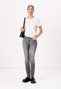 Camiseta gris claro con logo, jeans ajustados grises y zapatos negros gruesos. El modelo sostiene un pequeño bolso negro frente a un fondo simple.
