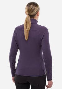 Femme avec une queue de cheval blonde portant une veste texturée à manches longues violette et un pantalon noir ajusté, vue de dos sur fond blanc.
