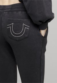 Czarne spodnie dresowe o miękkiej fakturze, z ozdobnym białym haftowanym logo na tylnej kieszeni oraz elastycznym pasem.