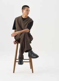 Brun stickad väst över en svart t-shirt, tillsammans med bruna vida byxor och svarta klumpiga skor, sittande på en träpall.