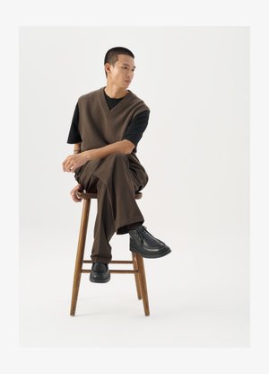 Barna kötött mellény fekete póló felett, barna bőszárú nadrággal és fekete vastag talpú cipővel párosítva, egy fa széken ül.