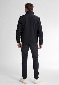 Giacca softshell nera con colletto alto, chiusura con zip e dettagli di cucitura, abbinata a jeans scuri e sneakers chiare.