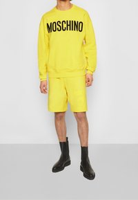 Osoba ubrana w jasny żółty sweter Moschino i dopasowane krótkie spodenki, w połączeniu z czarnymi botkami, stojąca na jednolitym tle.