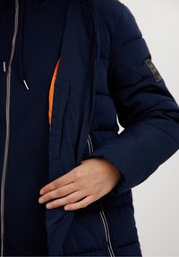 Námořnická puffer bunda s prošívanou strukturou a oranžovou kapsou; má zapínání na zip a kapuci se stahovací šňůrkou.
