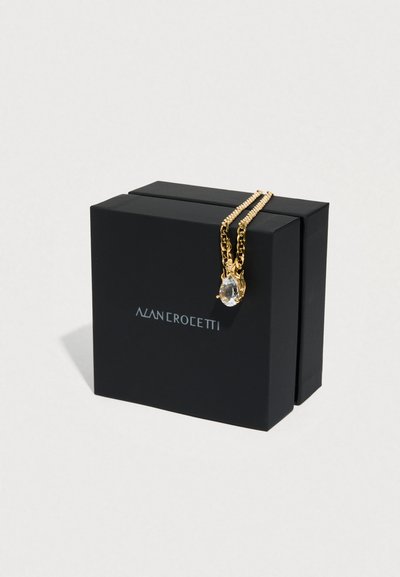 Χρυσό κολιέ με αλυσίδα που διαθέτει διαφανές μενταγιόν, τοποθετημένο πάνω σε μαύρο κουτί με το όνομα "ALAN CROCEETTI" τυπωμένο σε ασημί. Υφασμένες επιφάνειες και κομψός σχεδιασμός.