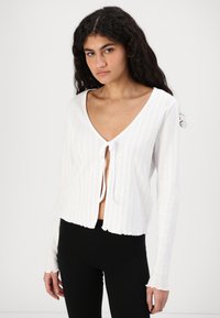 Bijela rebrasta cardigan jakna s dubokim V-izrezom, dugim rukavima i prednjim vezanjem. Ima valoviti rub i teksturni uzorak za dodatne detalje.