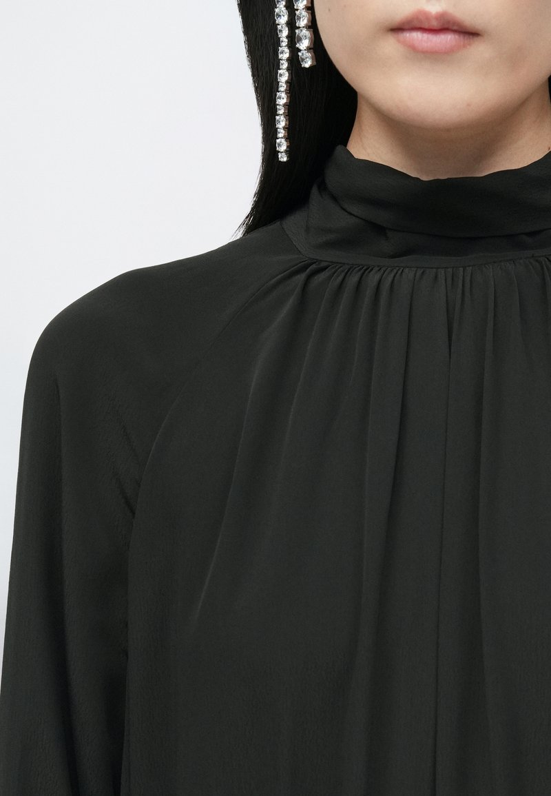Blusa negra de cuello alto en tejido ligero con pliegues y textura sutil, que presenta un pendiente decorativo de strass en el lado izquierdo.