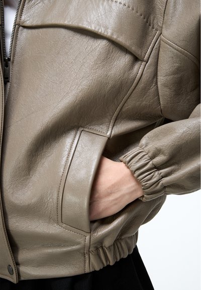 Taupefarbene Lederjacke mit elastischen Bündchen und Saum, Hand ruht in der vorderen Tasche, sichtbarer Reißverschluss und Druckknopfdetails.