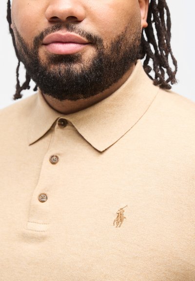 Άνδρας με γένια που φοράει μπεζ πόλο μπλουζάκι με γιακά και κουμπιά, με μικρό κ εντητό λογότυπο αλόγου και αναβάτη στο στήθος.