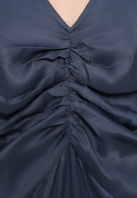 Rochie bleumarin cu decolteu în V, având pliuri strânse în față și un material neted, cu textură, care curge într-o siluetă în formă de A.